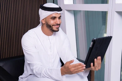 اجراء مكالمة فيديو عبر الانترنت، استخدام الاجهزة التقنية الحديثة، مفهوم إدارة المشاريع عن بعد، رجل عربي خليجي اماراتي يرتدي الكندورة والغترة يجلس مقابل واجهة زجاجية مطلة على المدينة يقوم بانجاز مهام العمل عبر جهاز التابلت