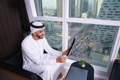 الاحترافية والتفاني في الإنجاز، استخدام الاجهزة التقنية الحديثة، مفهوم إدارة المشاريع عن بعد، رجل عربي خليجي اماراتي يرتدي الكندورة والغترة يجلس مقابل واجهة زجاجية مطلة على المدينة يقوم بانجاز مهام العمل عبر جهاز التابلت