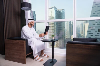 مفهوم إدارة المشاريع عن بعد، استخدام الاجهزة التقنية الحديثة، رجل عربي خليجي اماراتي يرتدي الكندورة والغترة يجلس مقابل واجهة زجاجية مطلة على المدينة يقوم بانجاز مهام العمل عبر جهاز التابلت، الاحترافية والتفاني في الإنجاز