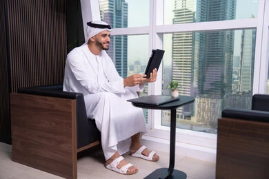 The use of modern technological devices, the concept of remote project management, an Arab Gulf Emirati man wearing a kandura and ghutrah sitting in front of a glass facade overlooking the city, completing work tasks via a tablet, professionalism and dedication in achievement.