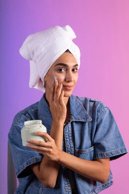 ترطيب الوجه بتقنيات التدليك اللطيف، الاهتمام بالمظهر الخارجي، مفهوم العناية بنضارة وتحسين البشرة، بورتريه مقرب لامرأة عربية خليجية سعودية ترتدي الكاجوال و  منشفة بيضاء تضع كريم مرطب، استخدام كريمات و مكونات تجميلية، خلفية ملونه