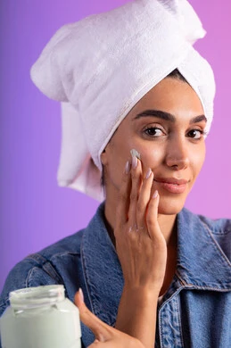 تدليك الوجه لنعومة وترطيب طبيعي، الاهتمام بالمظهر الخارجي، مفهوم العناية بنضارة وتحسين البشرة، بورتريه مقرب لامرأة عربية خليجية سعودية ترتدي الكاجوال و  منشفة بيضاء تضع كريم مرطب، استخدام كريمات و مكونات تجميلية، خلفية ملونه