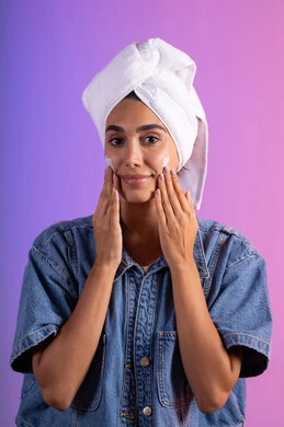 عناية وترطيب الوجه بالتدليك اللطيف، الاهتمام بالمظهر الخارجي، مفهوم العناية بنضارة وتحسين البشرة، بورتريه مقرب لامرأة عربية خليجية سعودية ترتدي الكاجوال و  منشفة بيضاء تضع كريم مرطب، استخدام كريمات و مكونات تجميلية، خلفية ملونه