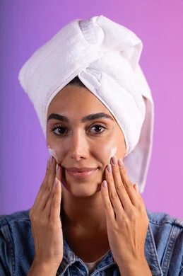 ترطيب وتنعيم الوجه بالتدليك، الاهتمام بالمظهر الخارجي، مفهوم العناية بنضارة وتحسين البشرة، بورتريه مقرب لامرأة عربية خليجية سعودية ترتدي الكاجوال و  منشفة بيضاء تضع كريم مرطب، استخدام كريمات و مكونات تجميلية، خلفية ملونه