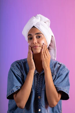 جلسة تدليك وترطيب للبشرة، الاهتمام بالمظهر الخارجي، مفهوم العناية بنضارة وتحسين البشرة، بورتريه مقرب لامرأة عربية خليجية سعودية ترتدي الكاجوال و  منشفة بيضاء تضع كريم مرطب، استخدام كريمات و مكونات تجميلية، خلفية ملونه