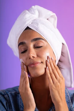 تدليك البشرة لزيادة الترطيب، الاهتمام بالمظهر الخارجي، مفهوم العناية بنضارة وتحسين البشرة، بورتريه مقرب لامرأة عربية خليجية سعودية ترتدي الكاجوال و  منشفة بيضاء تضع كريم مرطب، استخدام كريمات و مكونات تجميلية، خلفية ملونه