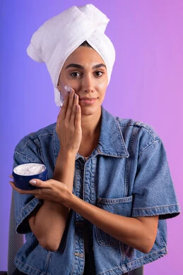 Facial care through massage and moisturizing, attention to external appearance, the concept of caring for the freshness and improvement of the skin, a close-up portrait of a Saudi Gulf Arab woman wearing casual attire and a white towel applying moisturizer, using creams and cosmetic ingredients, colorful background.