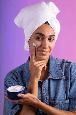 تدليك و ترطيب الوجه، الاهتمام بالمظهر الخارجي، مفهوم العناية بنضارة وتحسين البشرة، بورتريه مقرب لامرأة عربية خليجية سعودية ترتدي الكاجوال و  منشفة بيضاء تضع كريم مرطب، استخدام كريمات و مكونات تجميلية، خلفية ملونه