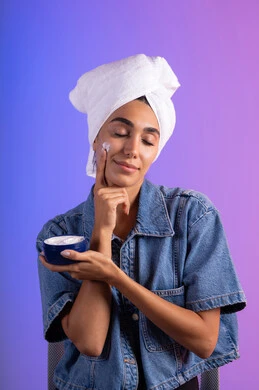 تدليك و ترطيب الوجه، الاهتمام بالمظهر الخارجي، مفهوم العناية بنضارة وتحسين البشرة، بورتريه مقرب لامرأة عربية خليجية سعودية ترتدي الكاجوال و  منشفة بيضاء تضع كريم مرطب، استخدام كريمات و مكونات تجميلية، خلفية ملونه