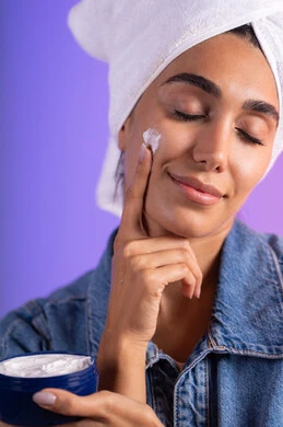 تدليك و ترطيب الوجه، الاهتمام بالمظهر الخارجي، مفهوم العناية بنضارة وتحسين البشرة، بورتريه مقرب لامرأة عربية خليجية سعودية ترتدي الكاجوال و  منشفة بيضاء تضع كريم مرطب، استخدام كريمات و مكونات تجميلية، خلفية ملونه