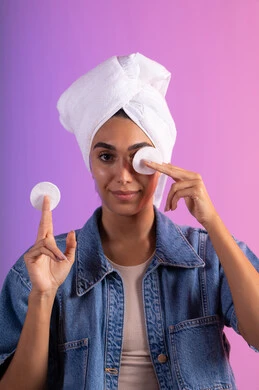 تنظيف البشرة من المكياج بالقطن، الاهتمام بالمظهر الخارجي، مفهوم العناية بنضارة ونظافة البشرة، بورتريه مقرب لامرأة عربية خليجية سعودية ترتدي الزي الكاجوال تضع منشفة بيضاء على رأسها تمسك بيديها قطنتين تقوم بمسح وجهها، خلفية ملونة