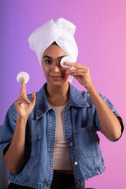 مسح المكياج باستعمال الضمادات القطنية، الاهتمام بالمظهر الخارجي، مفهوم العناية بنضارة ونظافة البشرة، بورتريه مقرب لامرأة عربية خليجية سعودية ترتدي الزي الكاجوال تضع منشفة بيضاء على رأسها تمسك بيديها قطنتين تقوم بمسح وجهها، خلفية ملونة