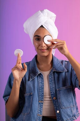 Removing makeup with cotton pads, caring about appearance, the concept of maintaining skin freshness and cleanliness, a close-up portrait of a Saudi Gulf Arab woman wearing casual attire, with a white towel on her head, holding cotton pads in her hands to wipe her face, colorful background.