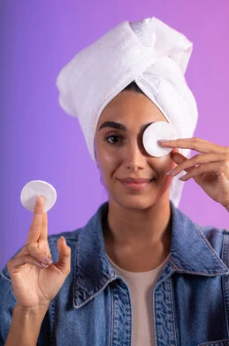 إزالة المكياج باستخدام ضمادات قطنية، الاهتمام بالمظهر الخارجي، مفهوم العناية بنضارة ونظافة البشرة، بورتريه مقرب لامرأة عربية خليجية سعودية ترتدي الزي الكاجوال تضع منشفة بيضاء على رأسها تمسك بيديها قطنتين تقوم بمسح وجهها، خلفية ملونة