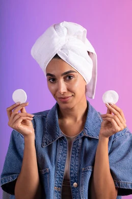 إزالة المكياج باستخدام ضمادات قطنية، مفهوم العناية بنضارة ونظافة البشرة، الاهتمام بالمظهر الخارجي، بورتريه لامرأة عربية خليجية سعودية ترتدي الزي الكاجوال تضع منشفة بيضاء على رأسها تمسك بيديها قطنتين، خلفية ملونة