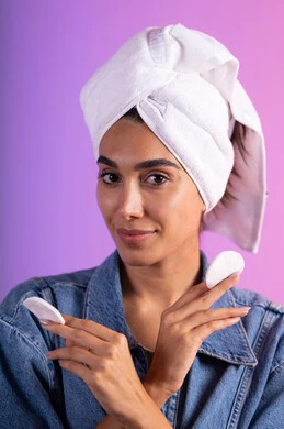 الاهتمام بالمظهر الخارجي، مفهوم العناية بنضارة ونظافة البشرة، بورتريه لامرأة عربية خليجية سعودية ترتدي الزي الكاجوال تضع منشفة بيضاء على رأسها تمسك بيديها قطنتين ، إزالة المكياج باستخدام ضمادات قطنية، خلفية ملونة