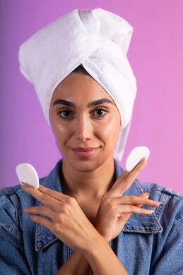 إزالة آثار المكياج باستخدام قطن الوجه، مفهوم العناية بنضارة ونظافة البشرة، الاهتمام بالمظهر الخارجي، بورتريه مقرب لامرأة عربية خليجية سعودية ترتدي الزي الكاجوال تضع منشفة بيضاء على رأسها تمسك بيديها قطنتين، خلفية ملونة