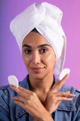 تنظيف الوجه من المكياج بالضمادات القطنية، مفهوم العناية بنضارة ونظافة البشرة، الاهتمام بالمظهر الخارجي، بورتريه مقرب لامرأة عربية خليجية سعودية ترتدي الزي الكاجوال تضع منشفة بيضاء على رأسها تمسك بيديها قطنتين، خلفية ملونة