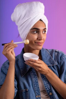 ايماءات السرور والسعادة، اتباع حمية غذائية، استخدام عسل النحل والمنتجات الطبيعية للعناية بالبشرة، منتج وطني محلي سعودي، بورتريه مقرب لامرأة عربية خليجية سعودية مبتسمة ترتدي الزي الكاجوال ومنشفة بيضاء تحمل بيدها ملعقة خشبية مغمورة بالعسل، خلفية ملونه