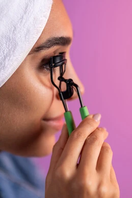 مفهوم الأنوثة والنعومة، بورترية مقرب من الجانب لإمرأة عربية خليجية سعودية ترتدي الزي الكاجوال تضع منشفة الرأس  تقوم  بالتزين ، استخدام أدوات تركيب الرموش، إبراز جمال العينين، الإهتمام بالجمال والمظهر الخارجي، خلفية ملونه