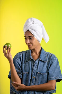 Enhancing skin freshness by consuming fresh fruits, following a healthy diet, and eating fresh fruit. A portrait of a young Arab Gulf Saudi woman wearing casual attire and a head towel, holding a green apple in her hand, with a colorful background.