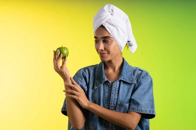 الحفاظ على اللياقة والوزن المثالي، اتباع نظام غذائي صحي وتناول الفاكهة الطازجة، بورتريه لشابة عربية خليجية سعودية ترتدي الزي الكاجوال و منشفة الرأس تحمل بيدها تفاحه خضراء، خلفية ملونه
