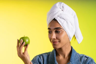 العناية بالجسم والحرص على الرشاقة، اتباع نظام غذائي صحي وتناول الفاكهة الطازجة، بورتريه لشابة عربية خليجية سعودية ترتدي الزي الكاجوال و منشفة الرأس تحمل بيدها تفاحه خضراء، خلفية ملونه