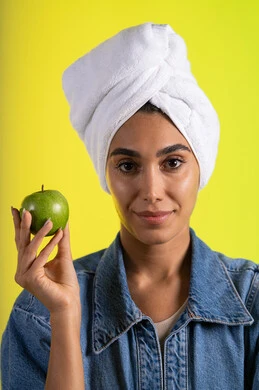 المحافظة على اللياقة البدنية والجسم المتناسق، اتباع نظام غذائي صحي وتناول الفاكهة الطازجة، بورتريه لشابة عربية خليجية سعودية ترتدي الزي الكاجوال و منشفة الرأس تحمل بيدها تفاحه خضراء، خلفية ملونه