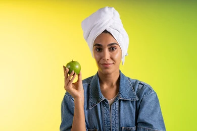 Attention to physical fitness, following a healthy diet and eating fresh fruit, a portrait of a young Arab Gulf Saudi woman wearing casual attire and a head towel holding a green apple in her hand, colorful background.