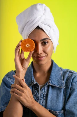 The role of natural fruits in skin freshness, meeting the body's need for nutrients, a close-up portrait of a Saudi Gulf Arab woman wearing casual attire with a white towel on her head holding slices of orange in her hands and placing them in front of her eyes, a delicious breakfast and snack, following a healthy diet, colorful background.