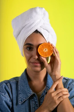 فوائد الفواكه الطازجة في تغذية البشرة، تلبية حاجة الجسم من العناصر الغذائية, بورتريه مقرب لامرأة عربية خليجية سعودية ترتدي الزي الكاجوال تضع منشفة بيضاء على رأسها تمسك  بيديها شرائح من البرتقال تضعها أمام عينها، فطور وسناك لذيذ، اتباع نظام غذائي صحي، خلفية ملونة