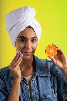 أهمية الفواكه الطازجة للبشرة، تلبية حاجة الجسم من العناصر الغذائية, بورتريه مقرب لامرأة عربية خليجية سعودية ترتدي الزي الكاجوال تضع منشفة بيضاء على رأسها تمسك بيديها شرائح من البرتقال و قطعه قطنتية، فطور وسناك لذيذ، اتباع نظام غذائي صحي، خلفية ملونة