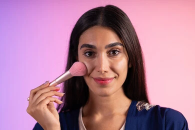 تحسين المظهر الخارجي والاعتناء بالجمال، التسويق لمنتجات التجميل، مكياج وميكاب تتوريال، صالون نسائي راقي، استخدام أدوات تجميل نسائيه عصريه، بورتريه مقرب لفتاه عربيه سعودية خليجيه ترتدي الزي الكاجوال تحمل بيدها فرشاة توريد الخدود، خلفية ملونه