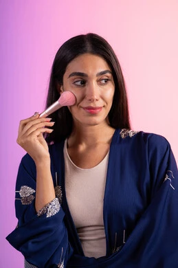 التسويق لمنتجات التجميل، مكياج وميكاب تتوريال، مفهوم الانوثه والاهتمام بالمظهر الخارجي، صالون نسائي راقي، استخدام أدوات تجميل نسائيه عصريه، بورتريه مقرب لفتاه عربيه سعودية خليجيه ترتدي الزي الكاجوال تحمل بيدها فرشاة توريد الخدود، خلفية ملونه