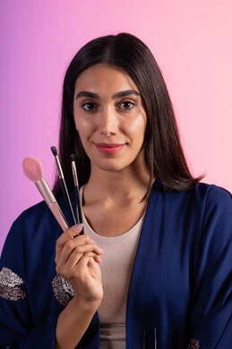 The concept of caring for beauty and skin, marketing cosmetic products, a store selling cosmetics, a close-up portrait of a Saudi Gulf Arab woman wearing casual attire holding a set of makeup brushes, makeup tutorial tools, colorful background.