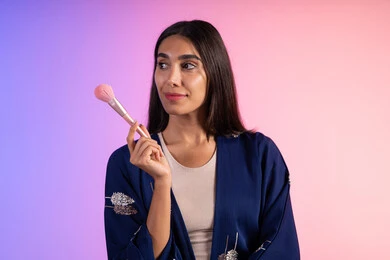 مكياج وميكاب تتوريال، مفهوم الانوثه والاهتمام بالمظهر الخارجي، صالون نسائي راقي، استخدام أدوات تجميل نسائيه عصريه، بورتريه مقرب لفتاه عربيه سعودية خليجيه ترتدي الزي الكاجوال تحمل بيدها فرشاة توريد الخدود، التسويق لمنتجات التجميل، خلفية ملونه