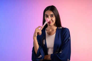 صالون نسائي راقي، استخدام أدوات تجميل نسائيه عصريه، بورتريه مقرب لفتاه عربيه سعودية خليجيه ترتدي الزي الكاجوال تحمل بيدها فرشاة توريد الخدود، مفهوم الانوثه والاهتمام بالمظهر الخارجي، مكياج وميكاب تتوريال، التسويق لمنتجات التجميل، خلفية ملونه