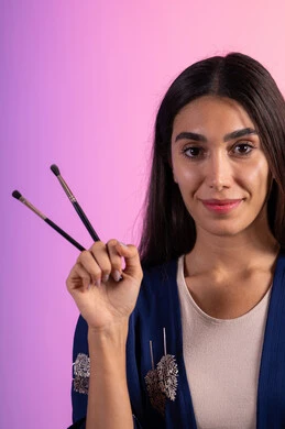 النظر الى الكاميرا بإيماءات النعومة والثقة، توزيع أحمر الخدود على الوجه بالفرشاة، الحفاظ على الجاذبية والمظهر الجميل، التسويق لأدوات ومنتجات التجميل, بورتريه مقرب لامرأة عربية خليجية سعودية ترتدي الزي الكاجوال تمسك فرش المكياج ، خلفية ملونة