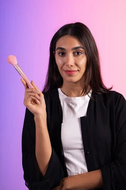 تطبيق أحمر الخدود باستخدام الفرشاة، الحفاظ على الجاذبية والمظهر الجميل، التسويق لأدوات ومنتجات التجميل, بورتريه مقرب لامرأة عربية خليجية سعودية ترتدي الزي الكاجوال تمسك فرش المكياج بإيماءات النعومة والثقة ، خلفية ملونة