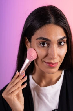 العناية بالبشرة بواسطة المستحضرات, مفهوم التزين والتجمل, الاهتمام بالمظهر الخارجي، بورتريه مقرب لامرأة عربية خليجية سعودية ترتدي الزي الكاجوال تستعمل فرشاة أحمر الخدود, خلفية ملونه