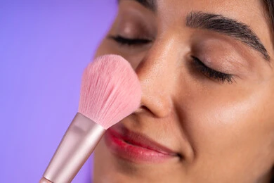 استعمال فرشاة أحمر الخدود، الحفاظ على الجاذبية والمظهر الجميل، التسويق لأدوات ومنتجات التجميل, بورتريه مقرب لامرأة عربية خليجية سعودية ترتدي الزي الكاجوال تمسك فرش المكياج بإيماءات النعومة والثقة ، خلفية ملونة