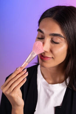 الحفاظ على الجاذبية والمظهر الجميل، التسويق لأدوات ومنتجات التجميل, بورتريه مقرب لامرأة عربية خليجية سعودية ترتدي الزي الكاجوال تمسك فرش المكياج بإيماءات النعومة والثقة, استعمال فرشاة أحمر الخدود ، خلفية ملونة