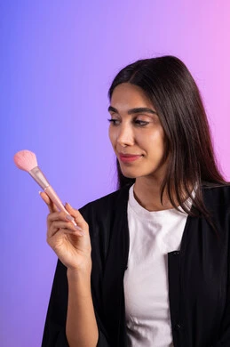 التزين والعناية بالمظهر الخارجي، التسويق لأدوات ومنتجات التجميل, بورتريه مقرب لامرأة عربية خليجية سعودية ترتدي الزي الكاجوال تمسك فرش المكياج بإيماءات النعومة والثقة, استعمال فرشاة أحمر الخدود ، خلفية ملونة