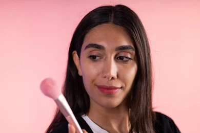 مفهوم تحسين المظهر والمكياج، التسويق لأدوات ومنتجات التجميل, بورتريه مقرب لامرأة عربية خليجية سعودية ترتدي الزي الكاجوال تمسك فرش المكياج بإيماءات النعومة والثقة, استعمال فرشاة أحمر الخدود ، خلفية ملونة