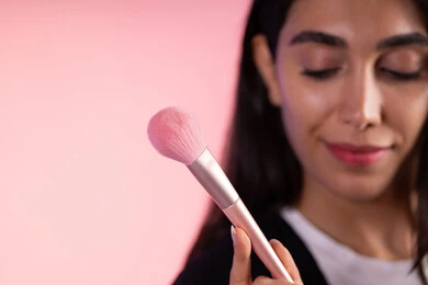 العناية بالجمال الخارجي والتزين، التسويق لأدوات ومنتجات التجميل, بورتريه مقرب لامرأة عربية خليجية سعودية ترتدي الزي الكاجوال تمسك فرش المكياج بإيماءات النعومة والثقة, استعمال فرشاة أحمر الخدود ، خلفية ملونة