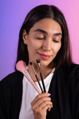 التسويق لأدوات ومنتجات التجميل, مفهوم التزين والتجمل, بورتريه مقرب لامرأة عربية خليجية سعودية ترتدي الزي الكاجوال تمسك فرش المكياج بإيماءات النعومة والثقة، الاهتمام والعناية بالمظهر الخارجي, استعمال فرشاة أحمر الخدود ، خلفية ملونة