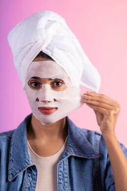 تطبيق مستحضرات تجميل للعناية الشخصية، روتين العناية وتحسين نضارة البشرة، خطوات تنظيف وتفتيح وترطيب الجلد, بورتريه مقرب لامراة عربية خليجية سعودية ترتدي الزي الكاجوال تضع منشفة الرأس تضع القناع التجميلي على الوجه، خلفية ملونه