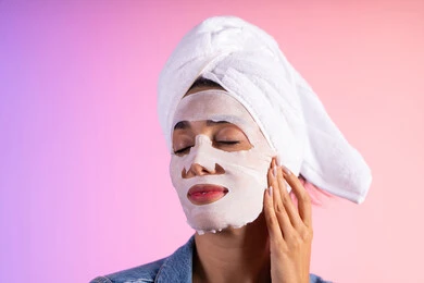 وضع مواد تجميلية للعناية بالمظهر، روتين العناية وتحسين نضارة البشرة، خطوات تنظيف وتفتيح وترطيب الجلد, بورتريه مقرب لامراة عربية خليجية سعودية ترتدي الزي الكاجوال تضع منشفة الرأس تضع القناع التجميلي على الوجه، خلفية ملونه