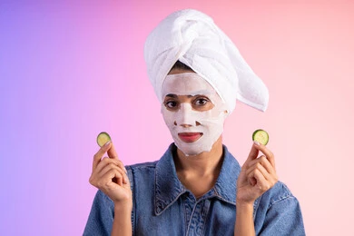 استعمال أدوات ومستحضرات التجميل ، مفهوم العناية والإهتمام بنضارة وتحسين البشرة ، بورتريه لمرأة عربية خليجية سعودية ترتدي الزي الكاجوال تضع منشفة الرأس تمسك بيديها قطع من الخيار، خلفية ملونه