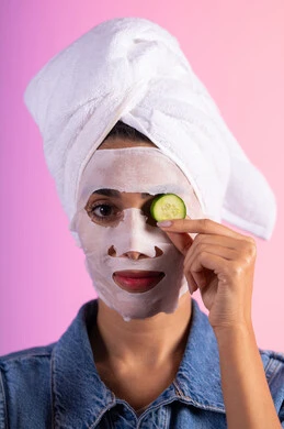 رعاية الجلد عبر التنظيف والترطيب والتفتيح، روتين العناية وتحسين نضارة البشرة، إستخدام مكونات تجميلية, بورتريه مقرب لامراة عربية خليجية سعودية ترتدي الزي الكاجوال و منشفة الرأس تضع القناع التجميلي على الوجه مع قناع الخيار للعيون، خلفية ملونه